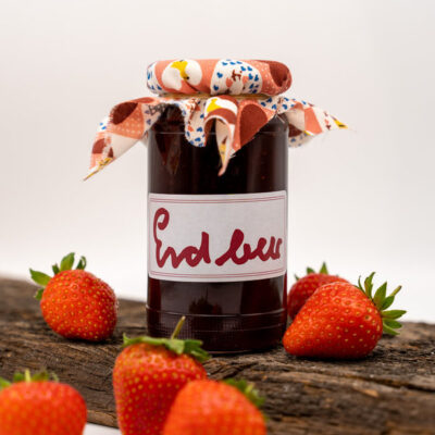 Glas Erdbeermarmelade mit Stoffabdeckung, umgeben von frischen Erdbeeren